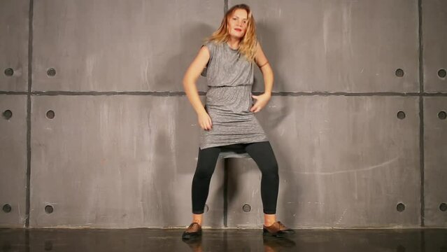 Blonde woman in gray dress originally dances near wall