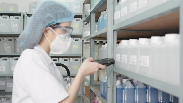 Beautiful young Asian chemist wears a hood, safety glasses using bar code reader to scanning bar code sticker on a plastic bottle containing a solvent on a shelf in a store to check information.