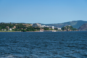 Fototapeta premium Distant view of Niteroi's Sao Domingos and Gragoata districts coastline as saw from Guanabara bay blue waters under summer afternoon sunny clear blue sky.