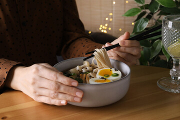 Woman eating delicious ramen with chopsticks at wooden table indoors, closeup. Noodle soup