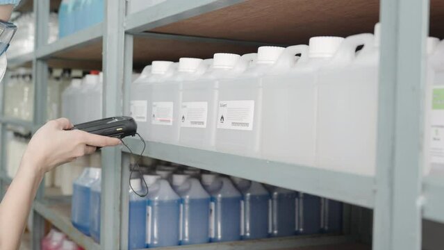 Beautiful young Asian chemist wears a hood, safety glasses using bar code reader to scanning bar code sticker on a plastic bottle containing a solvent on a shelf in a store to check information.