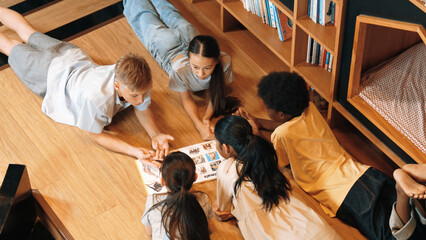 Group of diverse children lying down in circle while reading a book at library. Top view of girl sharing a magazine while pointing at interested topic and talking with lovely friends. Edification.