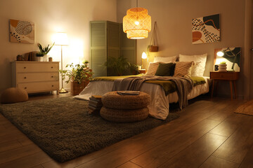 Interior of stylish bedroom with blankets on bed, wicker poufs and glowing lamps at night