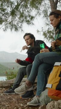 Vertical video. Traveling couple, a man and a woman, have stopped to have a snack while sitting on a log by the seaside. Strong wind and overcast weather. Mountains in the background covered with clou