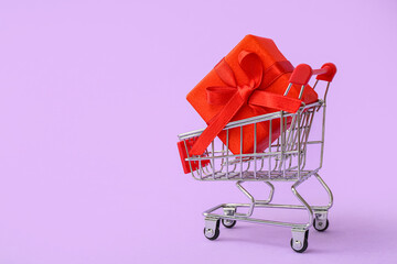 Small shopping cart with gift box on lilac background. Valentine's Day celebration