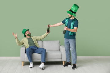 Young men in leprechaun hats with green beards holding bottles of beer near green wall. St....