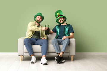 Happy young men in leprechaun hats with green beards holding bottles of beer and sitting on sofa...