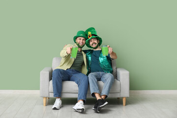Young men in leprechaun hats with green beards holding glasses of beer and sitting on sofa near...