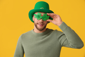 Young man in leprechaun hat and decorative glasses in shape of clover on yellow background. St. Patrick's Day celebration