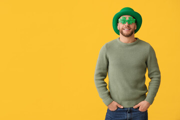 Happy young man in leprechaun hat and decorative glasses in shape of clover on yellow background....