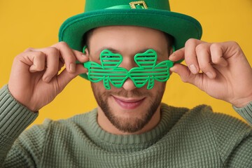 Young man in leprechaun hat and decorative glasses in shape of clover on yellow background. St....