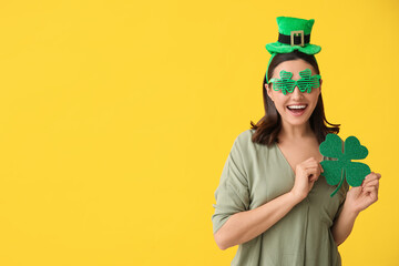 Beautiful young woman in leprechaun hat and decorative glasses with clover on yellow background....