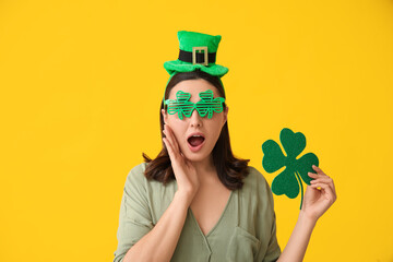 Shocked young woman in leprechaun hat and decorative glasses with clover on yellow background. St....