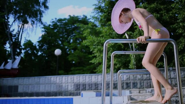 Young girl in swimwear and hat climbs by stairs on pool edge