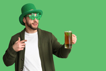 Young man in leprechaun hat and decorative glasses in shape of clover pointing at glass of beer on...