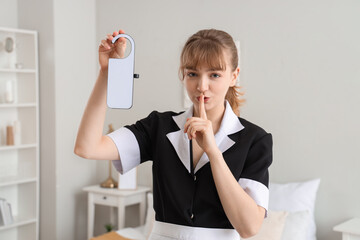 Young chambermaid with door hanger showing silence gesture in bedroom