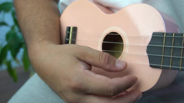 Man mid 30 and with bead is playing and whistling with his pink ukulele next to a big window and close to a green plant. Music is one form of art that can soothe the soul and make us feel better