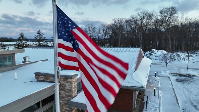 USA flag waving in winter snow scene in rural USA. Office building and forest woodland in distance.