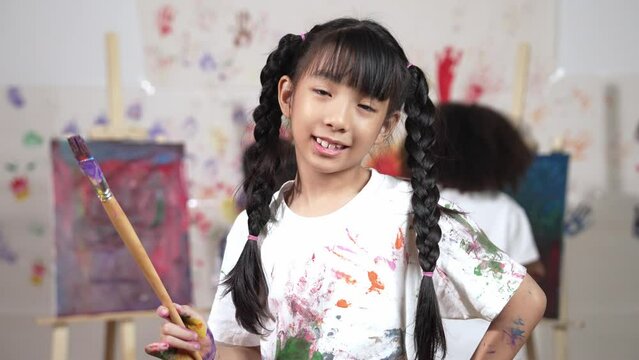Creative cute girl pose at camera while diverse children painting behind. Young beautiful child looking at camera while standing at stained room and holding paintbrush. Creative activity. Erudition.