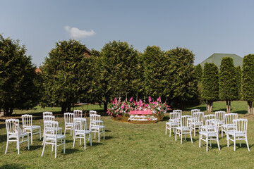 The pink stage is decorated with flowers on the background of the forest. Many white chairs on green grass. Preparation for the wedding ceremony