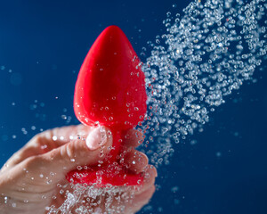Woman washing red anal plug under shower on blue background. 