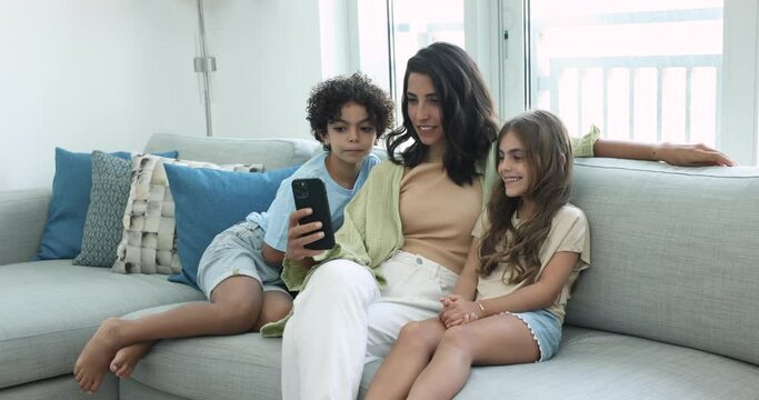 Happy Latin mom and two sibling son and daughter using smart home application on smartphone, enjoying online communication, relaxing together on couch, watching content, smiling, laughing, having fun