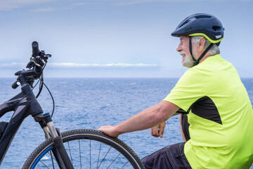 Happy active senior cyclist man enjoying riding with his bicycle at sea. Elderly bearded male with helmet looking at the horizon smiling. Active retirement lifestyle concept