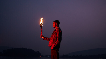 Image of brave man holding burning stick while moving in darkness. Young man holding a fire stick on the mountain top near the sea.  - Powered by Adobe