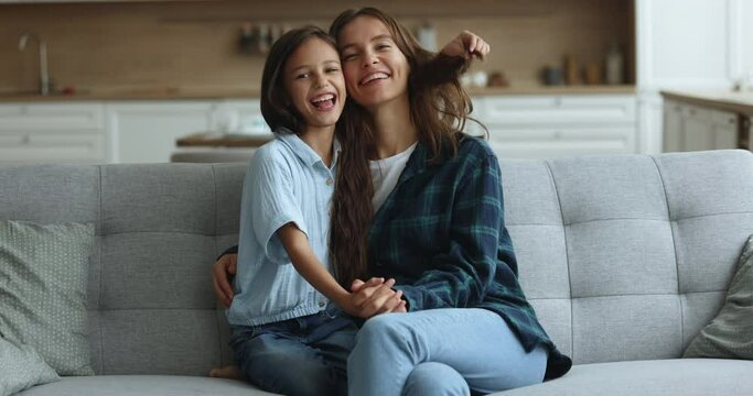 Cheerful beautiful young mom and cute kid girl hugging on cozy sofa at home, looking at camera with toothy smiles, laughing, holding hands, enjoying childhood, motherhood