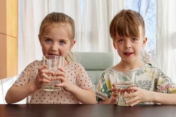 Brother and sister sit at kitchen table drinking water from glasses. Children quench thirst before going to school in morning closeup slow motion - Powered by Adobe