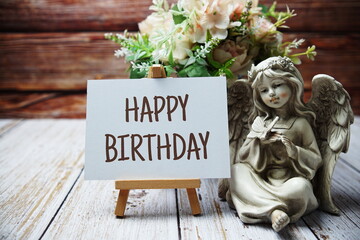 Happy Birthday text message written on paper card with wooden easel and angel statue with flower in...