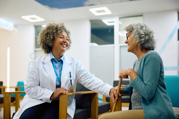 Happy African American doctor and her senior female patient talking in waiting room at medical clinic.