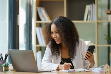 Portrait of a beautiful confident businesswoman using a laptop computer holding a mobile phone sitting in a modern office. Smiling African American freelancer working online from home.