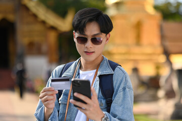 A handsome Asian tourist using a credit card and smartphone for cashless payment online for travel,...