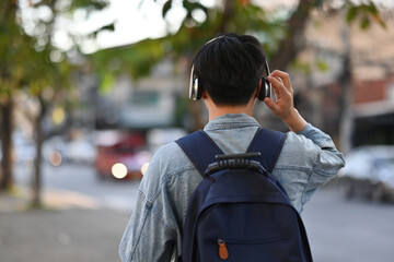 Rear view of Asian backpacker male walking on footpath while listening a music from headphones in the urban city, Solo travel concept