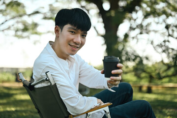 Smiling young Asian man sitting on camping chair handing a takeaway paper cup of coffee to you,...