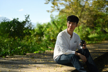 Obraz premium Portrait of a Young Asian man sitting on a wooden bench, enjoying a coffee and using a smartphone, outdoors with beautiful natural view