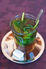A glass cup with Berber mint tea and metal saucer with a cube of sugar aside, traditional Moroccan hot beverage consumed over the hot days as a refreshing non-alcoholic drink