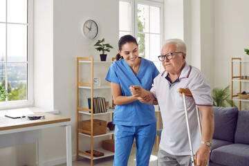 Friendly female nurse helping an injured senior man walk with a crutch, holding his hand, supporting him. Treatment and rehabilitation after injury, assisted living facility, senior care concept