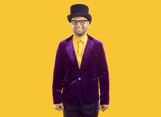 Studio shot of a male entertainer dressed for a theatrical performance or a circus show. Happy, smiling man in a purple velvet suit, top hat and eyeglasses standing isolated on a yellow background