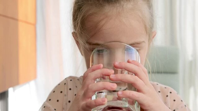 Pretty little girl drinking water from a glass in her hands in the kitchen at home, food and drink concept, indoor close-up portrait