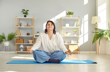 Fototapeta na wymiar Overweight woman doing yoga and meditation. Happy young fat girl sitting crossed legged on floor at home, doing exercises, breathing, relaxing, and meditating. Health, wellness, weight loss concept