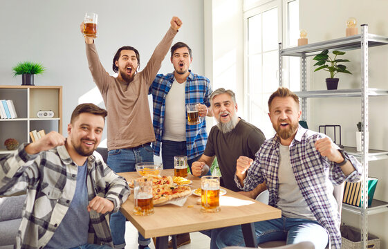 Group of male friends watching football at home. Several men enjoying weekend, having party, sitting at table in living room, eating food, drinking beer, watching soccer match, and shouting yay hooray