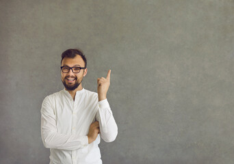 Got an idea. Happy positive young man in cool spectacles with forefinger pointing up standing on grey concrete copy space studio background. Cheerful guy thinks of good idea and points first finger up