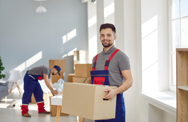Portrait of happy worker from professional man and van moving delivery service. Young male loader in uniform workwear standing in living room, holding cardboard box, looking at camera and smiling