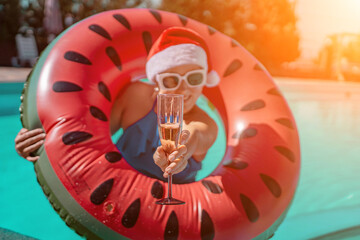 Woman pool Santa hat. A happy woman in a blue bikini, a red and white Santa hat and sunglasses poses near the pool with a glass of champagne standing nearby. Christmas holidays concept.
