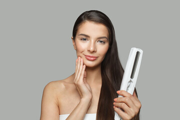 Cheerful model woman holding hair iron and straightening her healthy long dark hair on white...