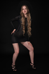 Fototapeta na wymiar Full length of young woman with long legs, posing on black background. Blonde model with long hair is wearing black short knitted transparent tight dress, wide brimmed felt hat and high heeled shoes