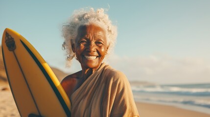 Fototapeta premium Happy elderly African surfer standing on the beach on a bright summer day. Smiling African grandmother standing on the shore with a surfboard. Cheerful old grandmother ready to surf the waves.