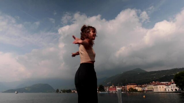 Young cute girl with curly hair is standing on the rocks at the edge of Lake Iseo. The girl has her back turned and she moves her arms simulating the wings of the plane